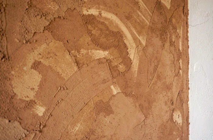 Mur intérieur recouvert d'un enduit en terre crue aux teintes ocre et sable, avec texture naturelle et légères imperfections artisanales